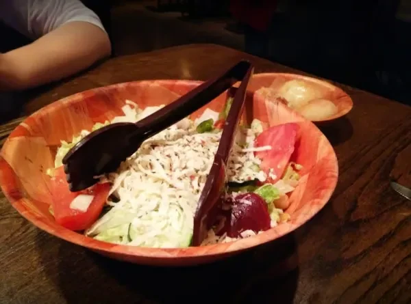 Dimly lit large wooden bowl of salad, tongs, rolls, person.