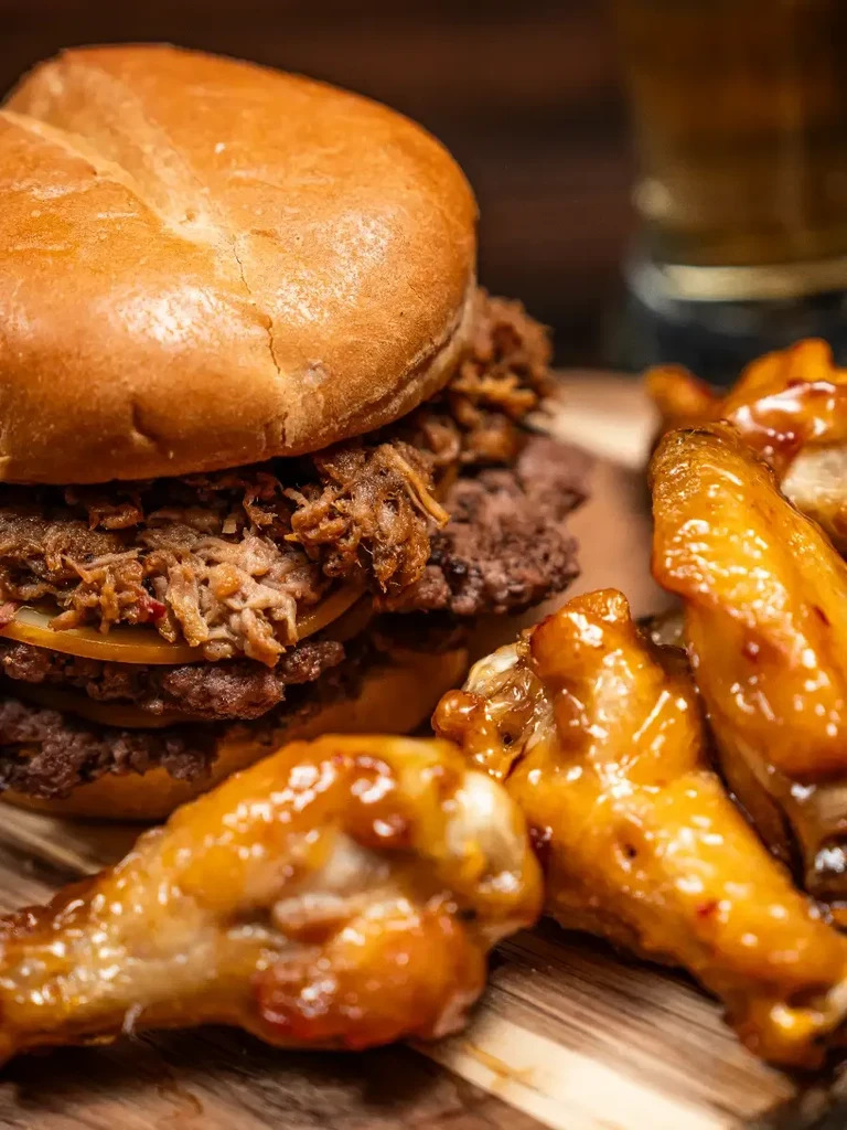 Close-up of glazed chicken wings and large burger on wooden board.