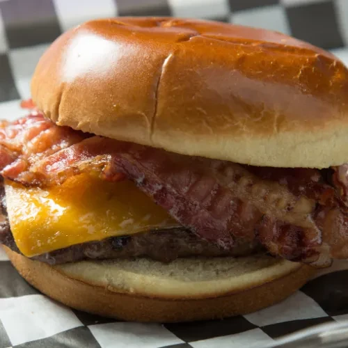 Close-up bacon cheeseburger with melted cheese and crispy bacon on checkered paper.