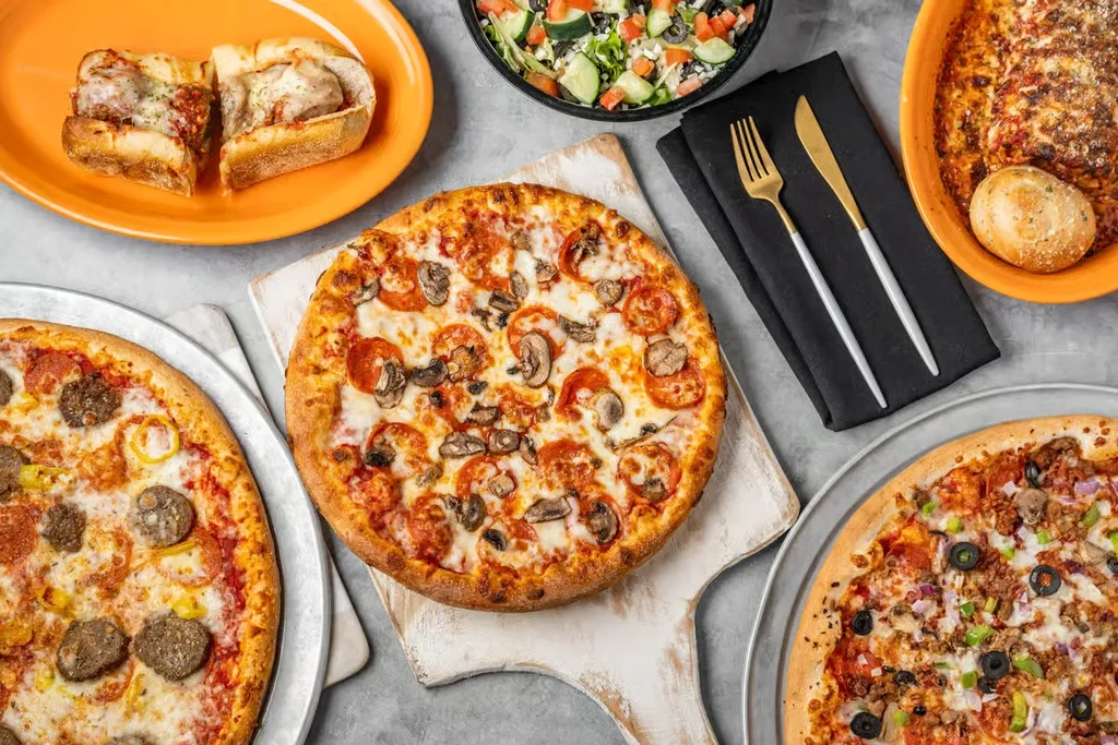 An overhead spread of various Italian-American pizzas, pasta, subs, and salad.