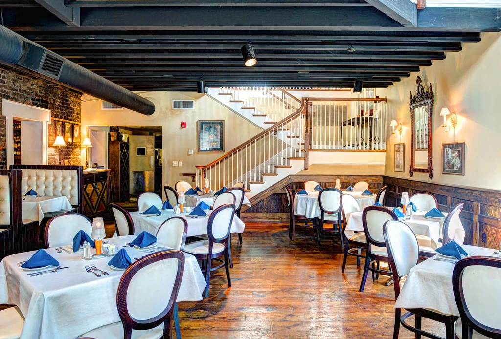 Empty restaurant dining room with set tables, elegant staircase, and brick walls.
