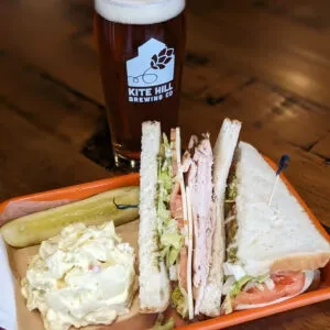 Deli sandwich, potato salad, pickle, and Kite Hill beer.