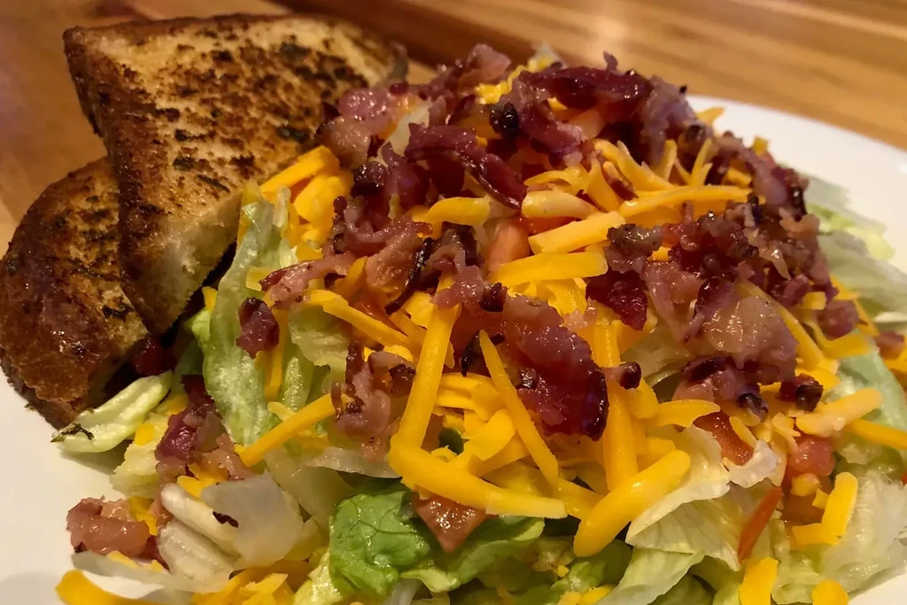 Salad with shredded cheddar, bacon, and grilled bread on a white plate.