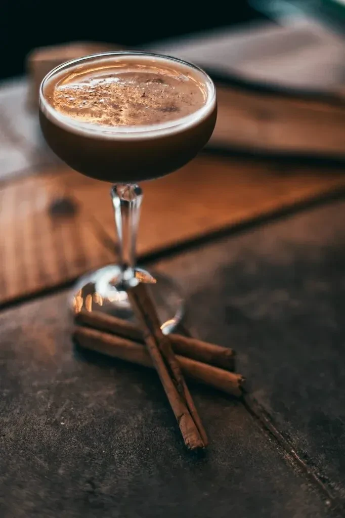 Frothy dark drink in coupe glass, garnished with three cinnamon sticks.