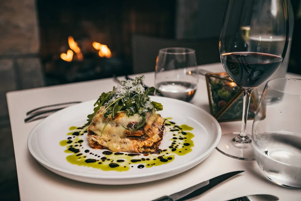 Plated baked pasta with cheese, arugula, wine, and blurred fireplace.