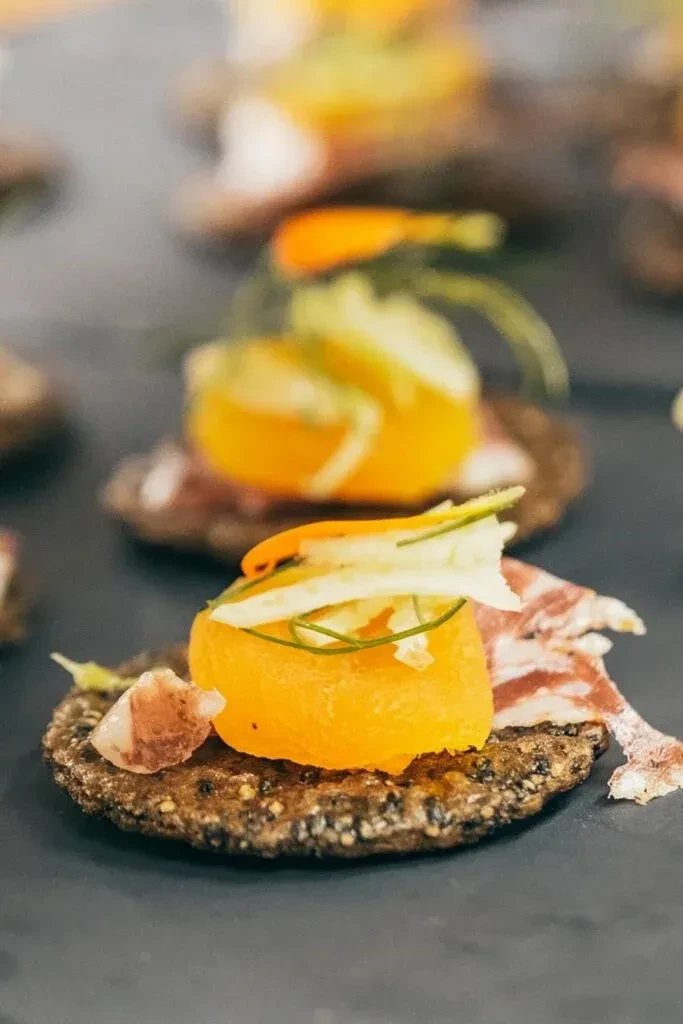 Elegant canapés with orange puree, vegetables, and cured meat on dark plate.