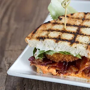 Grilled sandwich with bacon, fried patty, lettuce, and sauces on a plate.