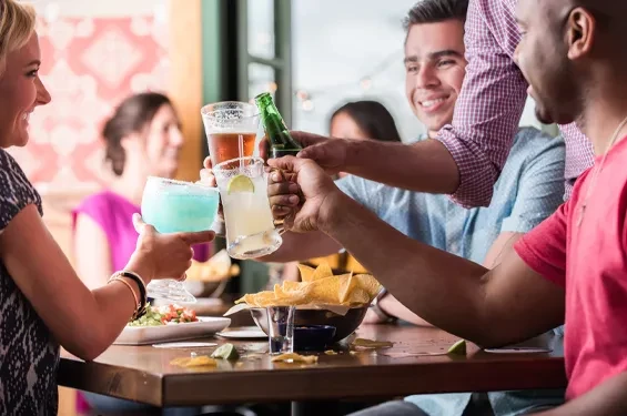 Friends' hands clinking various drinks over chips and salsa.