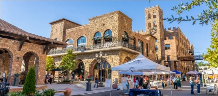 People browse an outdoor market near European buildings with a clock tower.