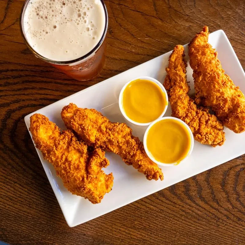 Golden-brown fried chicken tenders, yellow sauce, and beer on wood.