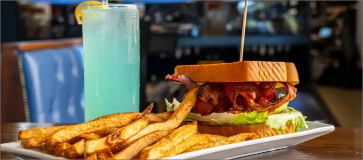 Bacon sandwich, golden fries, and turquoise cocktail on a wooden table.