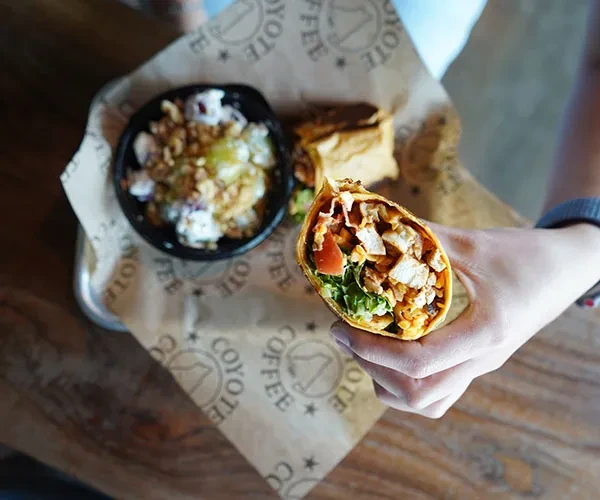 Overhead: Hand holding chicken wrap, side dish, on "Coyote Coffee" tray.