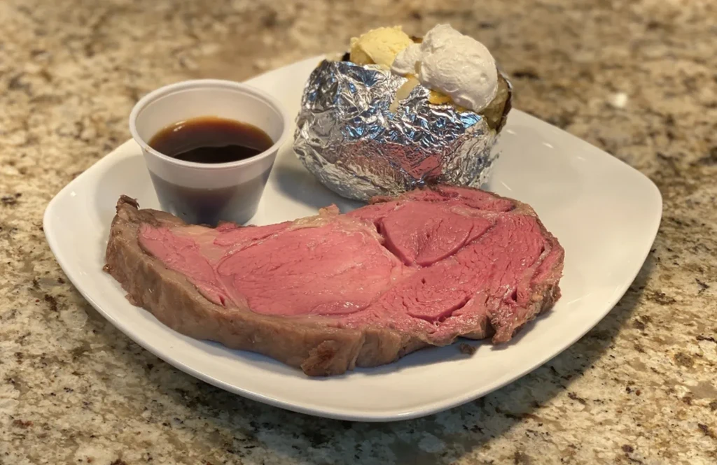 Medium-rare prime rib, baked potato, and au jus on a plate.