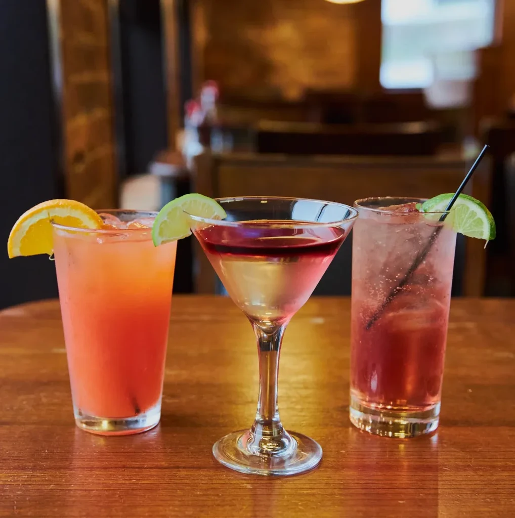 Three colorful cocktails on a wooden table, blurred bar background.