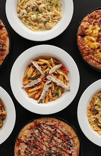 Top-down view of three pasta bowls and two pizzas on a dark table.