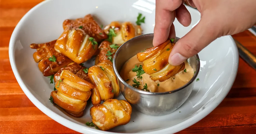 Hand dips herb-garnished pretzel bite into creamy sauce in a white bowl.