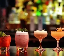 Four vibrant, garnished cocktails on a reflective bar with blurred lights.