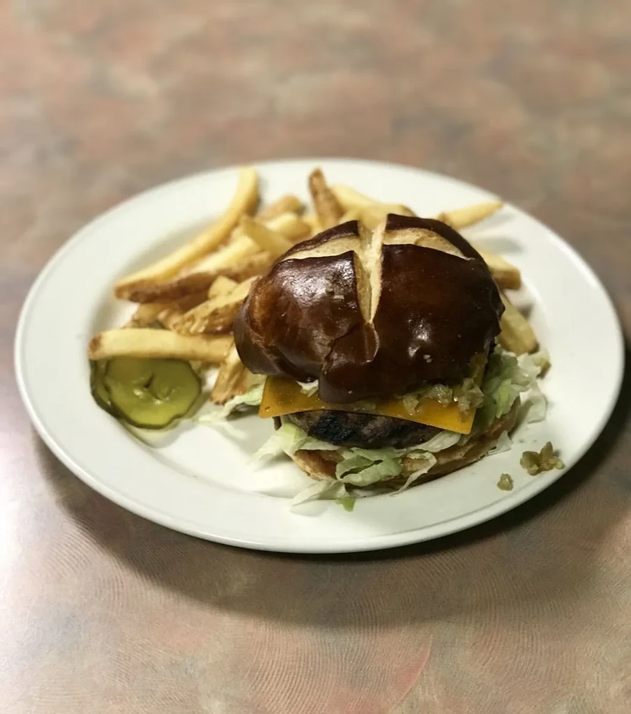 Pretzel bun burger with melted cheese, lettuce, fries, and pickle.