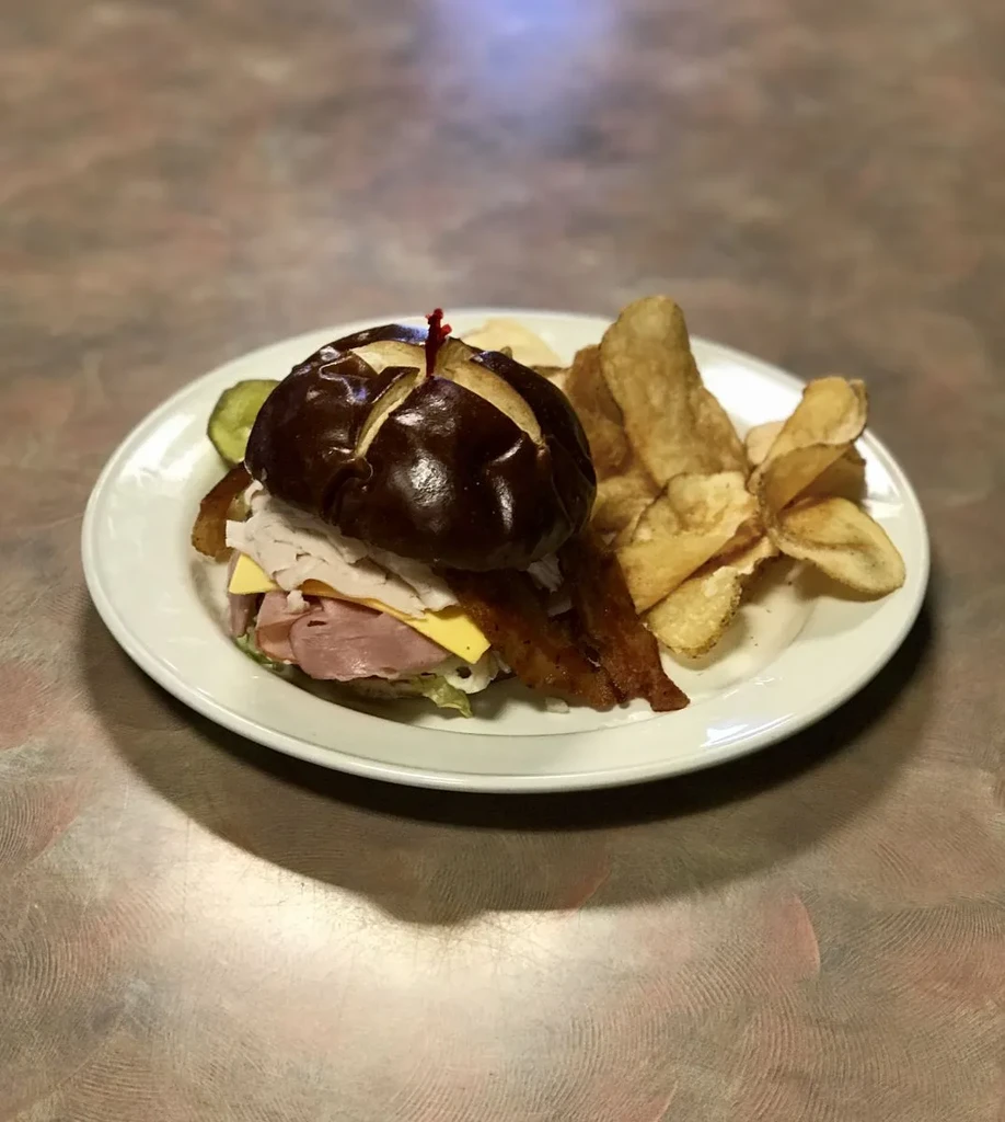 Deli meat sandwich on pretzel bun with chips and pickle.
