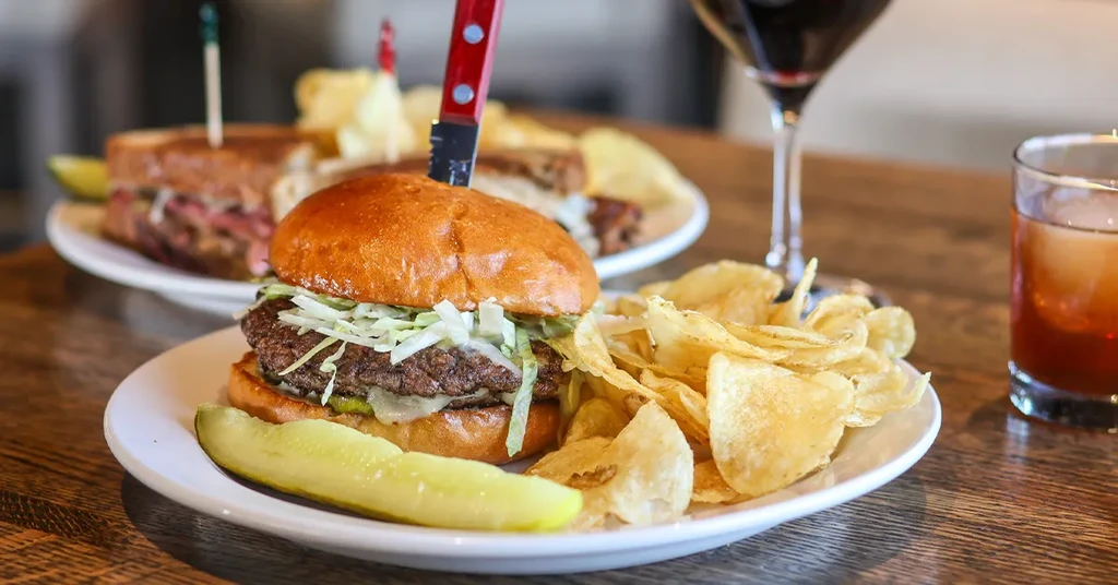 A hamburger with a knife, chips, and pickle on a wooden table.
