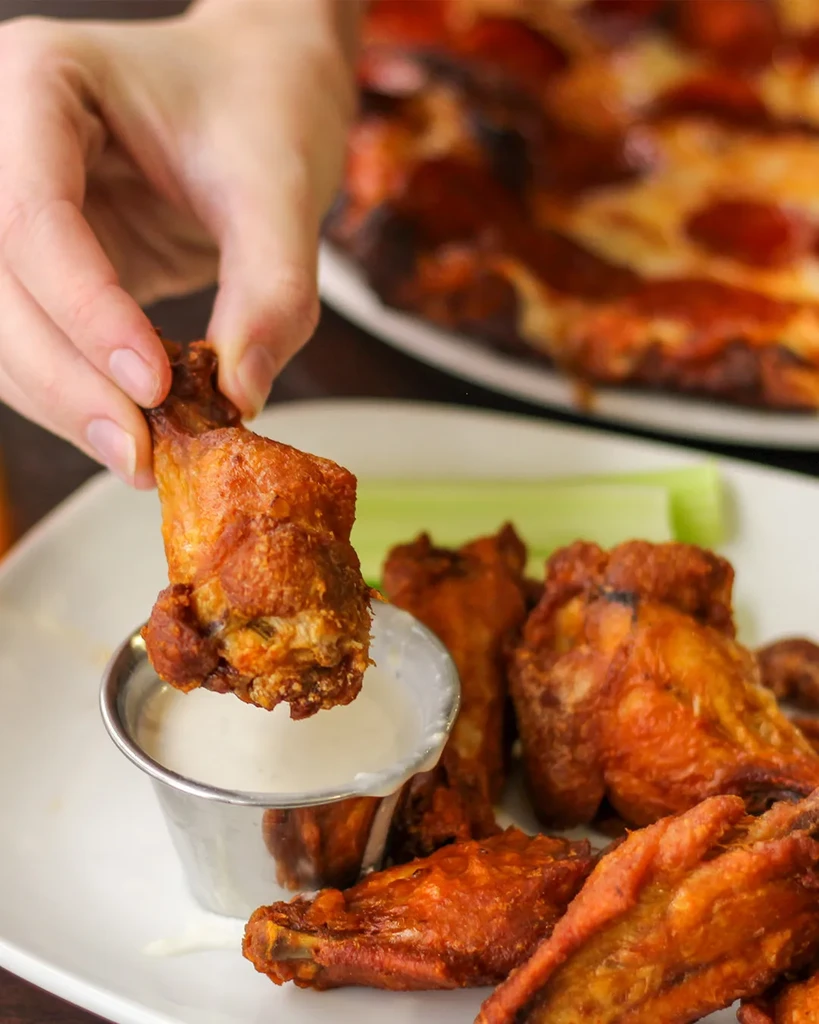 Hand dipping a crispy chicken wing into creamy white sauce.