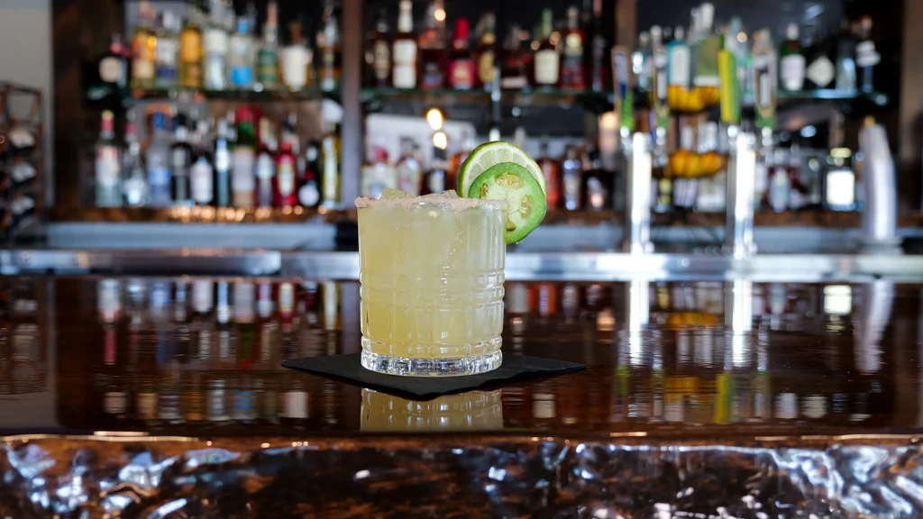 Pale yellow-green cocktail with lime, jalapeño, and rim at a bar.