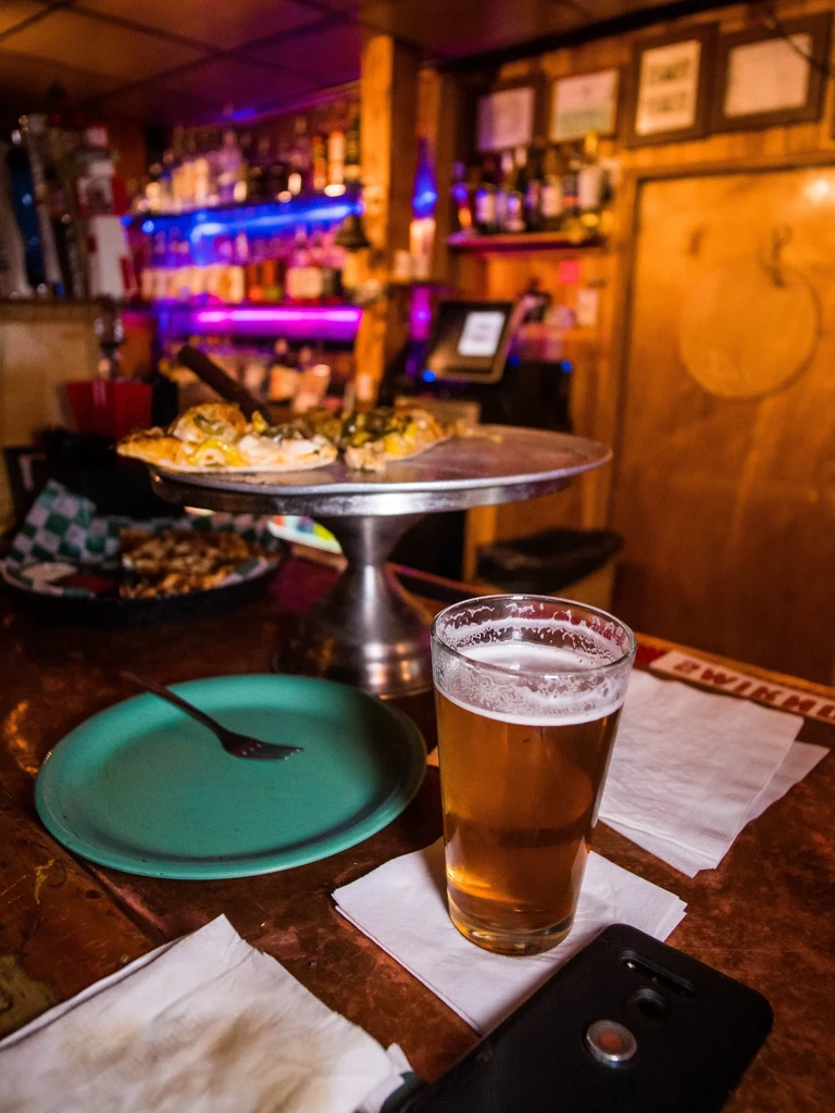 Rustic bar table with beer, plate, pizza, and colorful neon lights.