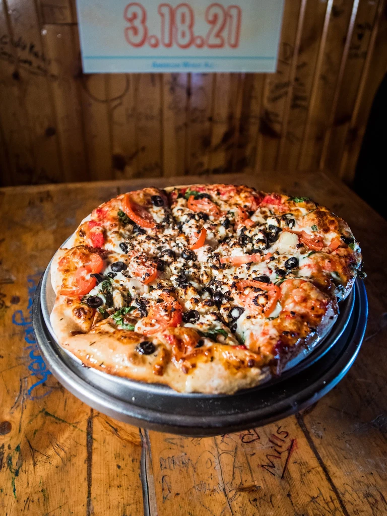 Generously topped pizza with tomatoes, olives, cheese on a rustic wooden table.