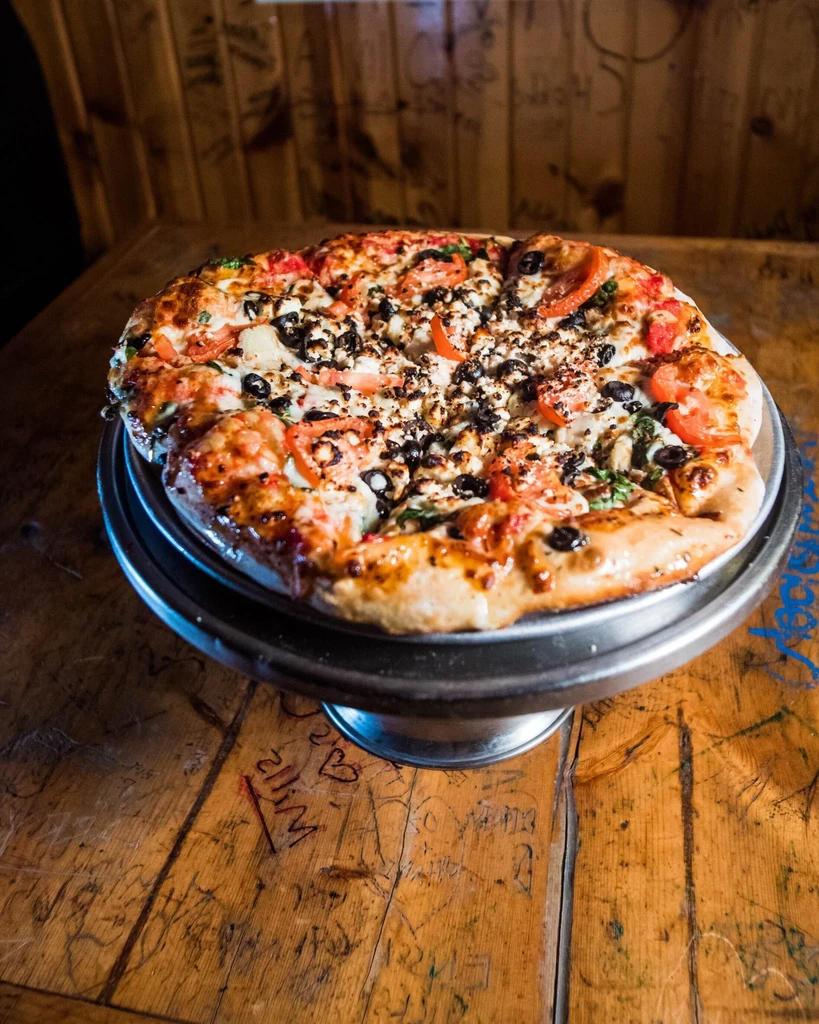 Pizza with tomato, olives, feta, and greens on a rustic carved table.