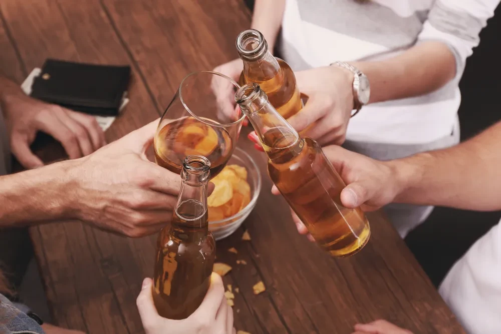 Multiple hands toast beer bottles and a glass with chips.