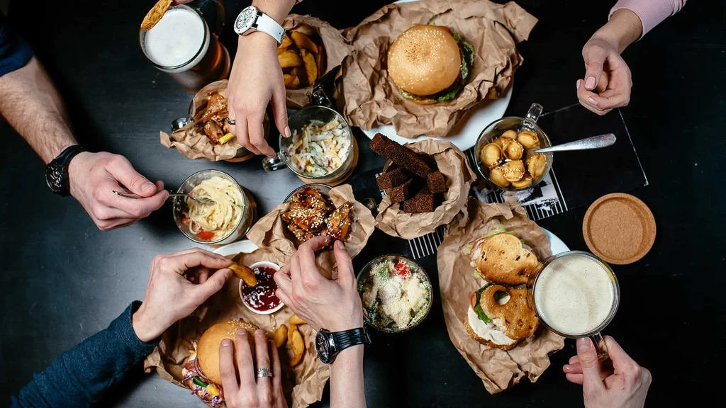 Overhead: Hands reach for burgers, fries, pasta, and beer on a table.