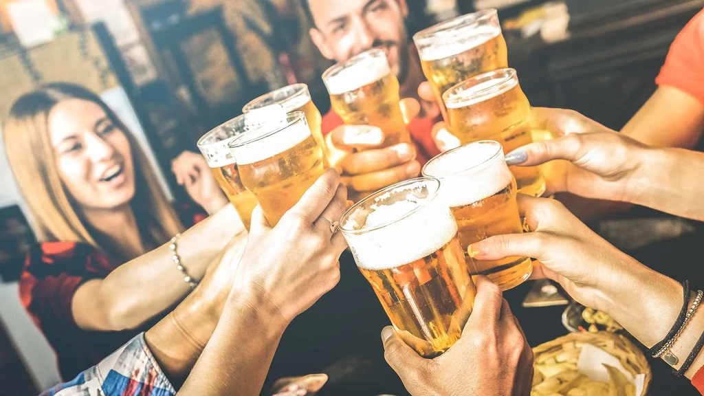 Hands toast with frothy beer glasses; blurred people behind.