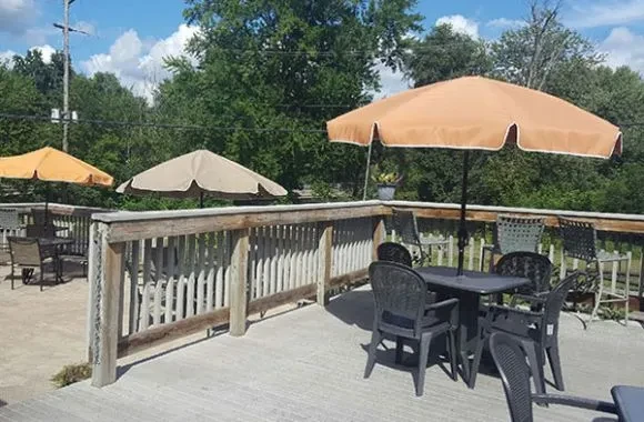 Sunny outdoor dining patio with tables, chairs, umbrellas, and green trees.