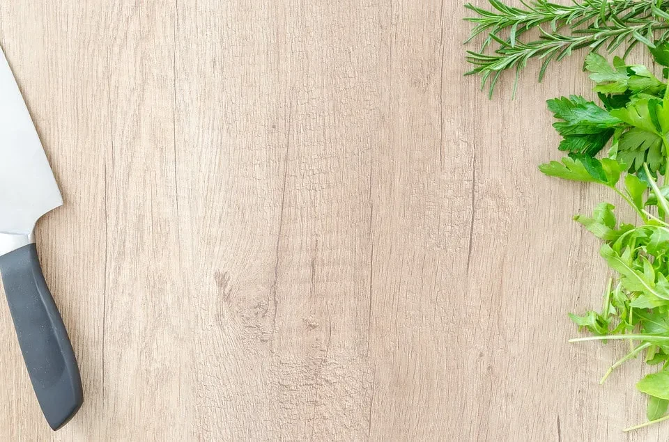 Chef's knife and fresh green herbs on a light wooden board.