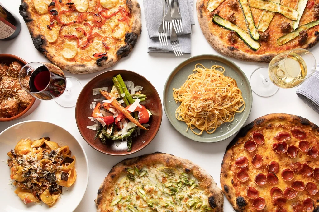 Overhead flat lay of Italian pizzas, pastas, meatballs, vegetables, and wine.