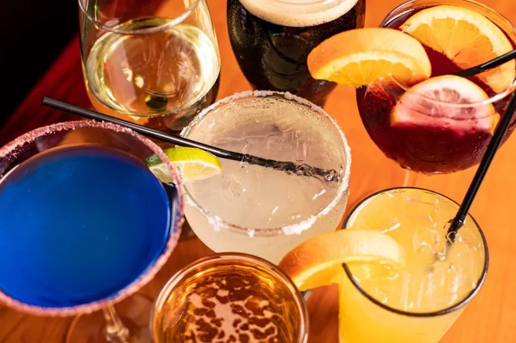 Top-down: Seven assorted beverages and cocktails on an orange surface.