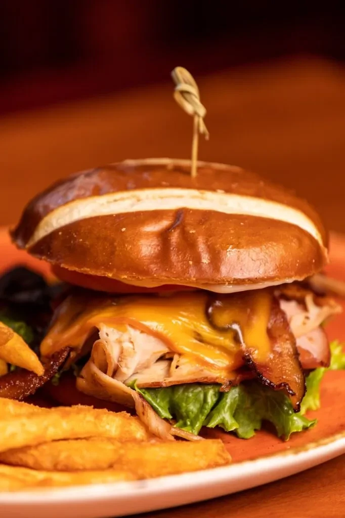 Close-up of a pretzel bun sandwich with turkey, bacon, cheese, and fries.
