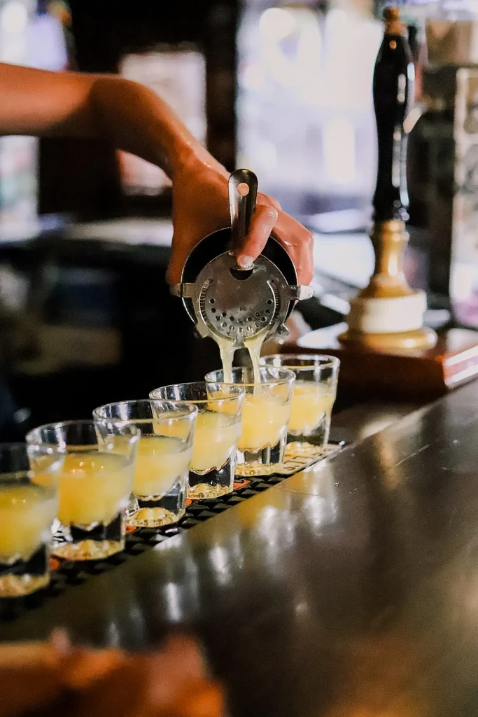 Bartender's hand pours cocktail from shaker into five shot glasses.