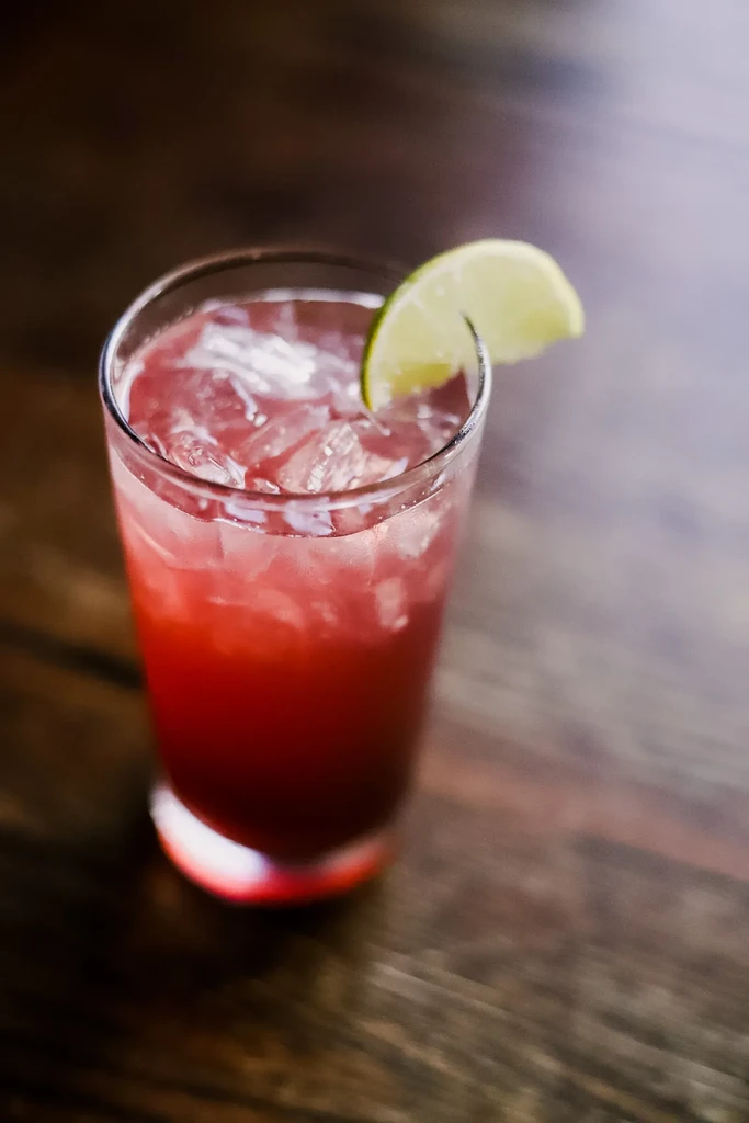 Reddish-pink drink with ice and lime wedge on dark wood.