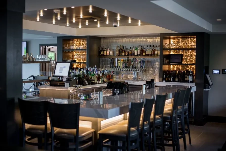 Empty modern bar, fully stocked with bottles, glassware, and stools.