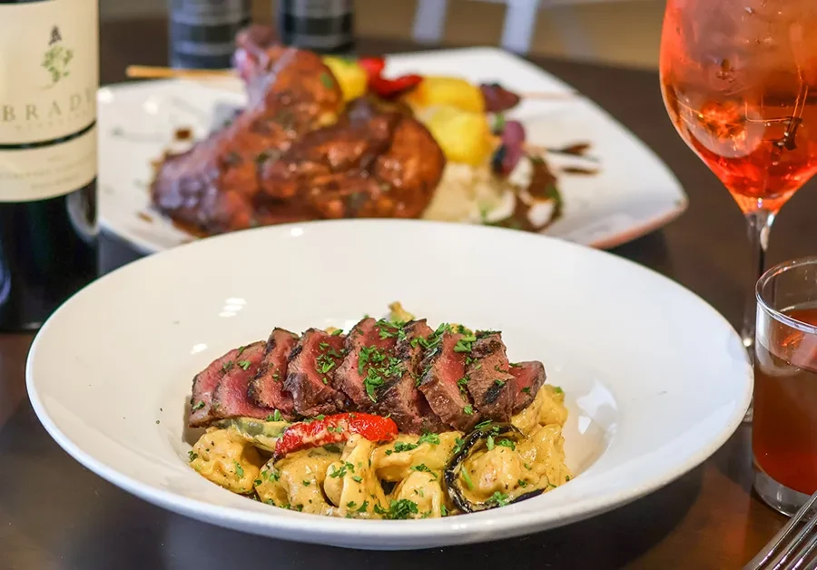 Close-up of seared steak and pasta dish, colorful skewers, wine, and cocktails.
