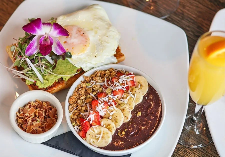 Elegant breakfast spread: avocado toast, acai bowl, bacon, orange drink.