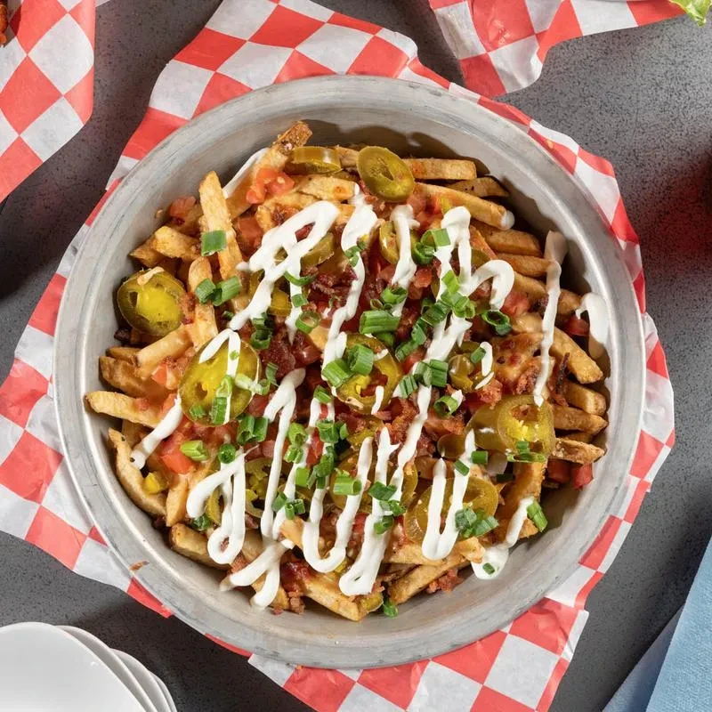Loaded french fries in metal bowl with jalapeños, bacon, tomatoes, sauce.