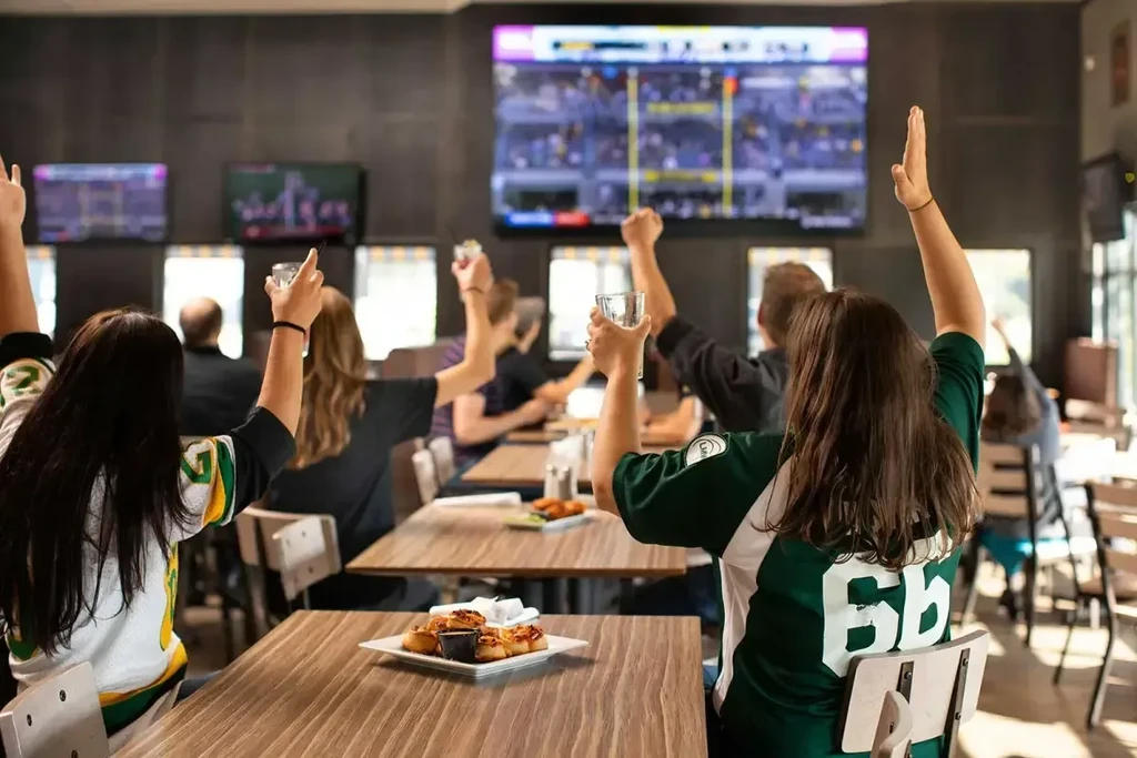 Patrons in a lively sports bar watching a game, food and drinks.