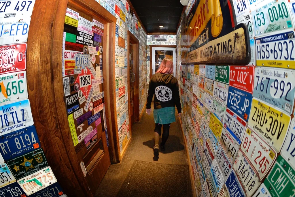Bar hallway walls covered in license plates, person with pink hair.