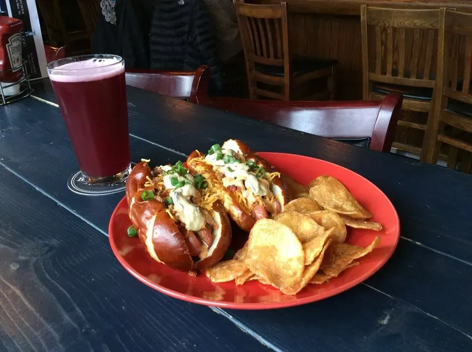 Two loaded hot dogs, chips, and a purple drink on wooden table.