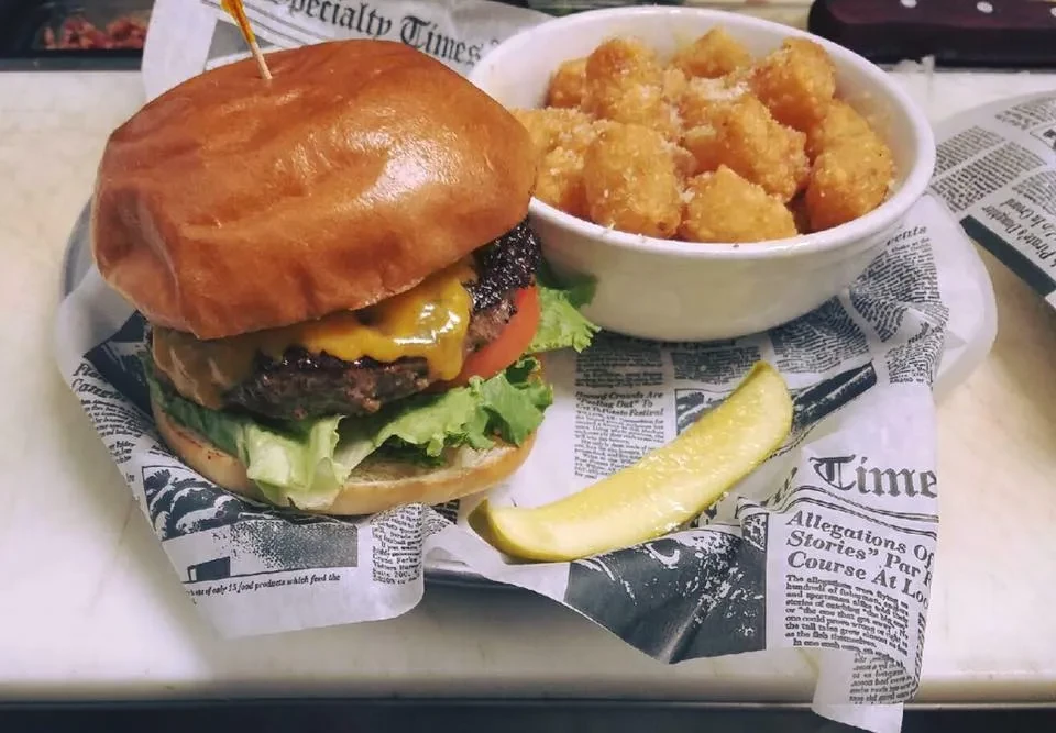 Cheeseburger, crispy tater tots, and pickle spear on newspaper-lined tray.