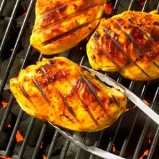 Tongs turning one of three golden-brown chicken breasts over coals.