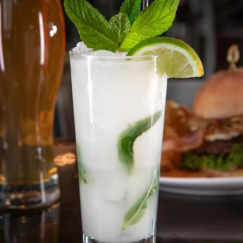 Cloudy drink with mint, lime, ice, and a straw, with food nearby.