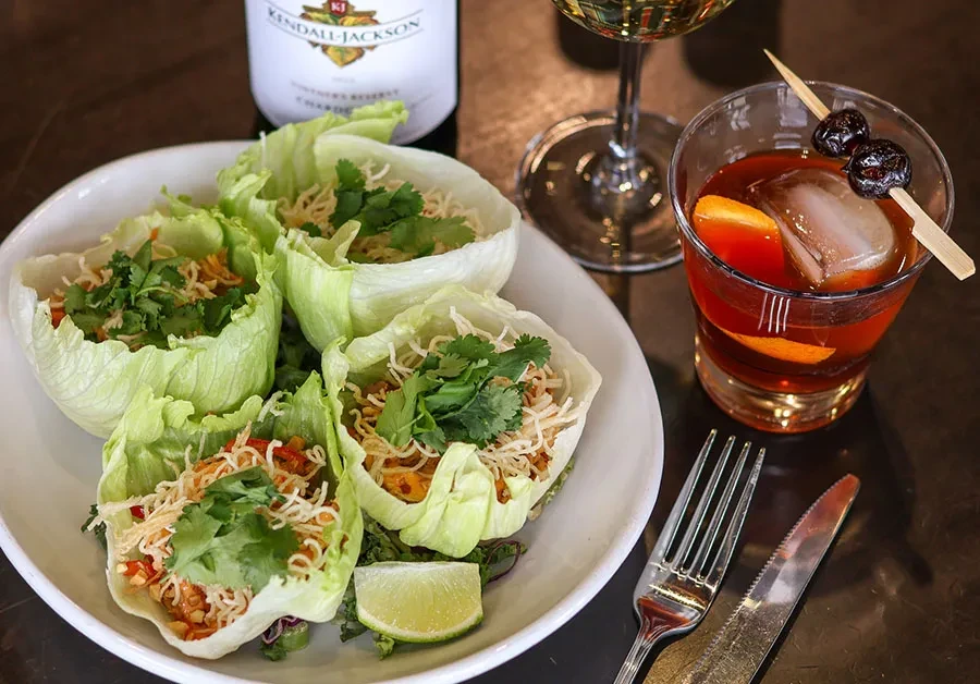 Four lettuce wraps with noodles, a cocktail, and Chardonnay wine on a table.